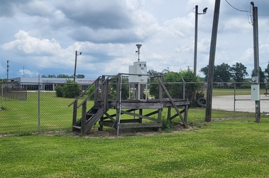 A wooden platform, accessible by stairs, that holds an FRM air sampling instrument in the middle of a grass field with a chain-link fence and trees running along the background.