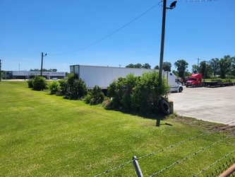 A grass field to the left with shrubs bordering it from a paved parking lot. There are large trucks and electrical poles in the lot, and trees are in the distance under a clear blue sky.