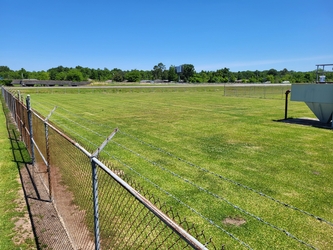 A grass field bordered by chain-link fencing in the left foreground with trees in the distant background.