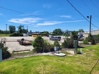 A chain-link fence running horizontally across the middle with grass in the foreground and a parking lot with vehicles behind the fence. There is a clear blue sky and trees in the distance.