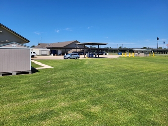 A grassy yard with a shed to the left and a large building in the background with cars parked under an overhang within a parking lot.