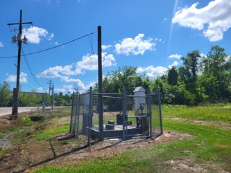 A small fenced-in area containing air monitoring equipment within a grass and dirt field with electrical lines and a partly cloudy blue sky overhead.