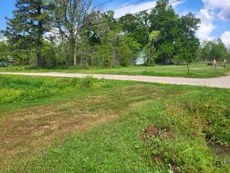 A grass field with a dirt vehicle path extending from the left and meeting perpendicularly with a gravel road; grass and large trees fill the background.