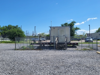 A small white shed on the bed of a trailer inside a fenced-in section of a gravel parking lot with cars on pavement in the background along with distant trees under a blue sky.