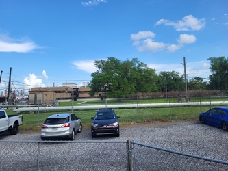 A gravel parking lot containing cars and surrounded by chain-link fencing, grass beyond the fence, and manmade structures and trees in the distance under a blue sky.