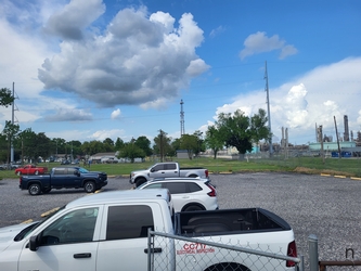 A parking lot filling most of the view with vehicles in the foreground and grass in the background, along with trees in the distance and a cloudy blue sky overhead.