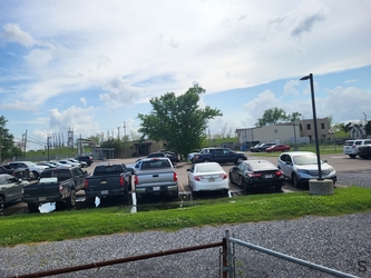 A gravel plot just behind chain-link fencing in the foreground, a strip of grass after the gravel plot, and then a parking lot filling the background with numerous cars.
