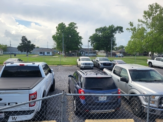 A fenced-in parking lot filled with cars in the foreground and residential homes lining the background with grass yards in between, trees scattered about, and a cloudy sky overhead.