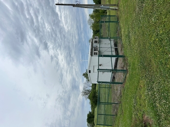 A grass field with a small shed inside a chain-link fenced-in square with more grass and trees in the background under a cloudy sky.