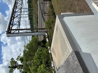 Part of the top of a small building in the foreground, trees beyond the building to the left, a fenced-off grass area to the right, and a highway in the background along with metal structures.