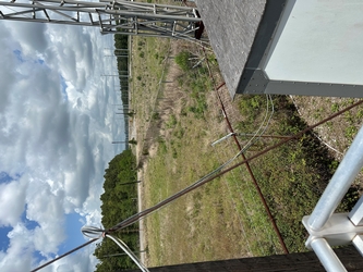 An expansive grassy field, fenced off into sections, with part of a building and metal railing in the foreground and a road with trees and clouds in the background.
