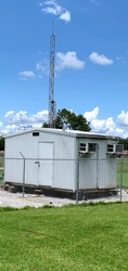 A small building in the center of a fenced-in section of a wide grassy field. A tall metal Met tower extends from the top of the building.