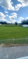 A vast grass field with trees in the background and part of a house visible towards the right. Some gravel and chain-link fencing are in the foreground.