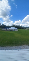 A vast grass field with trees in the far background, a residential home nearby, and fenced-in gravel in the foreground with part of a shed roof visible directly below.