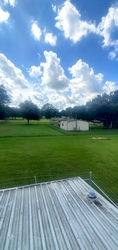 An expansive grass field with part of a shed roof visible in the foreground directly below. There is a house towards the middle of the property and trees filling the background.