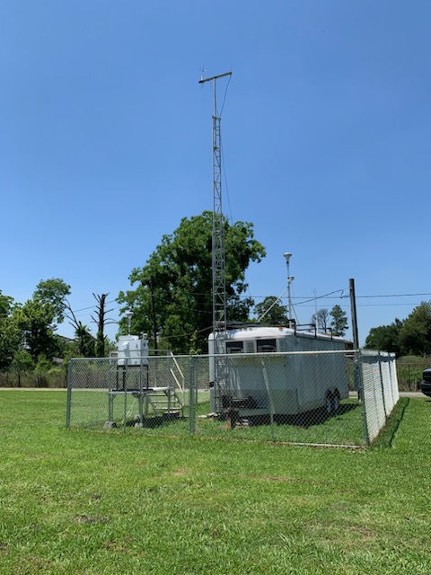A grassy piece of land with an enclosed metal trailer surrounded by a fence, a wooden platform holding air monitoring equipment to the left and a blue sky and trees in the background.