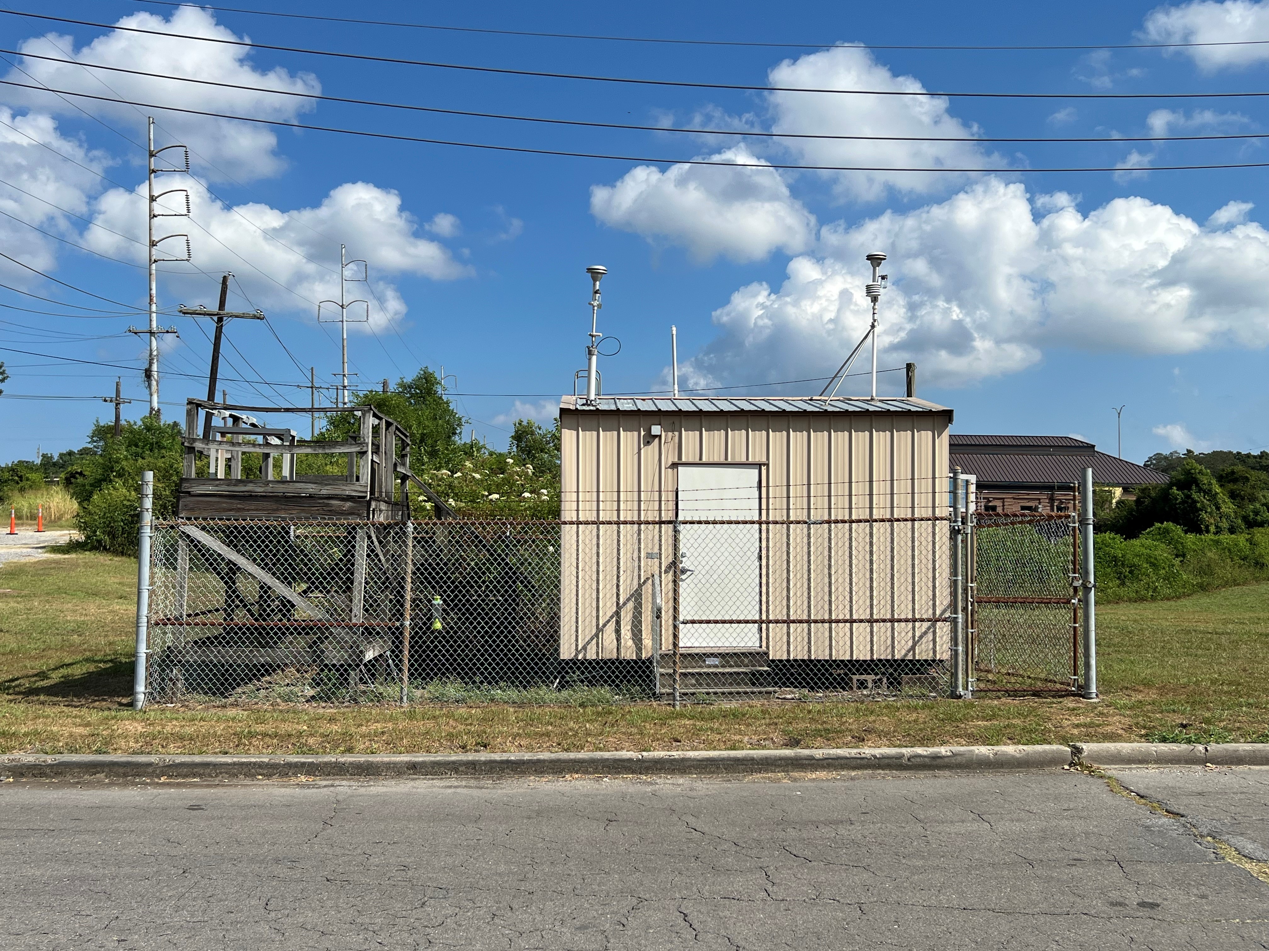 A tan metal shed inside a grassy, fenced in area with a street in the foreground and a wooden platform holding air monitoring equipment to the left.