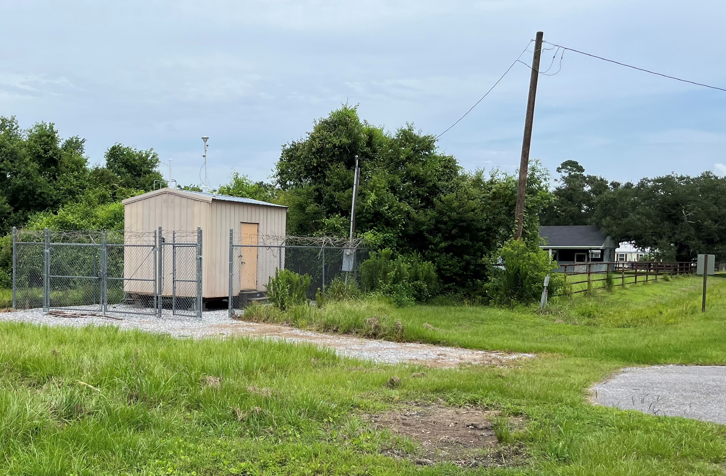 A small metal building with a gravel drive leading up to it through grass with trees and a house in the background.