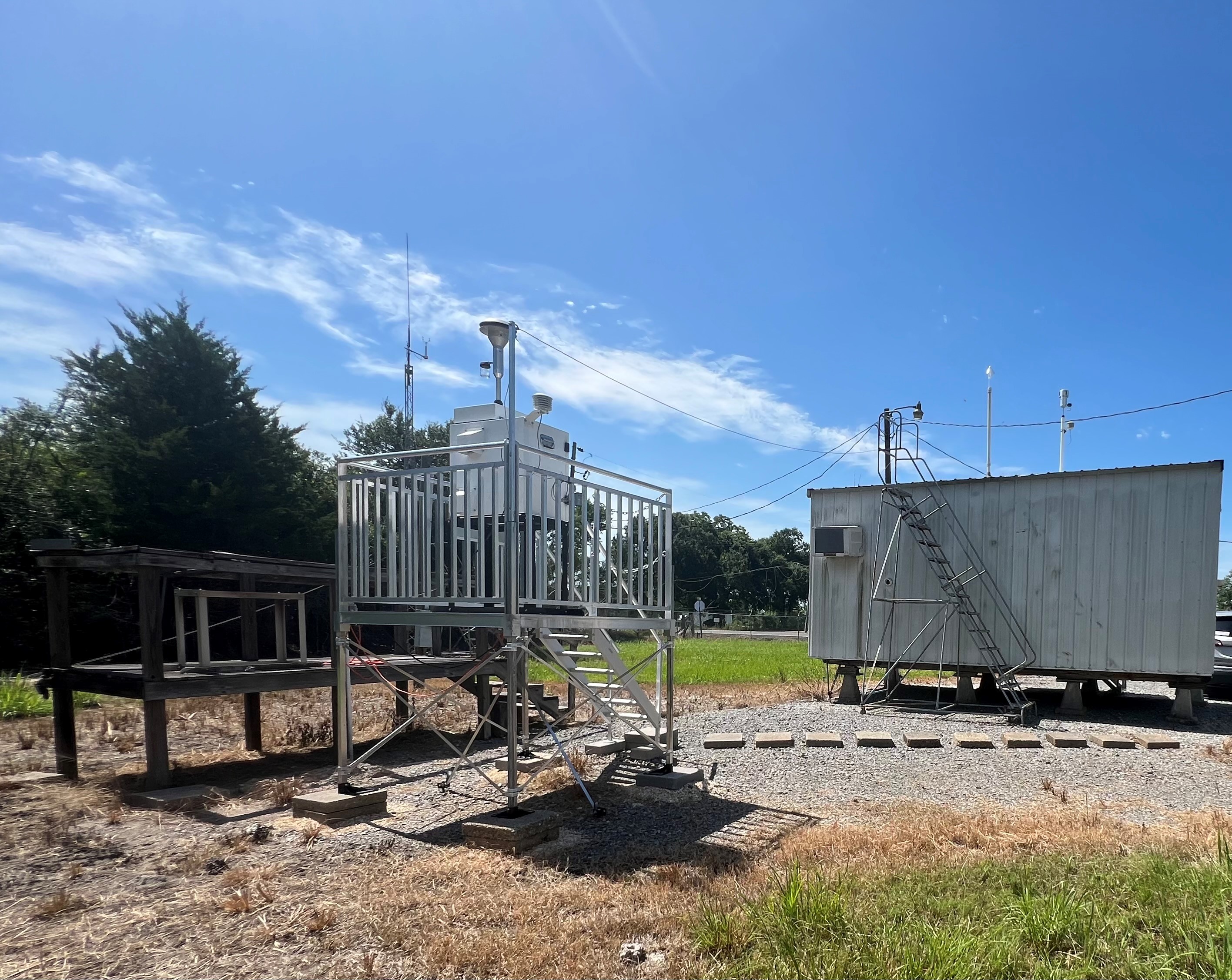 A metal platform with stairs holding an air monitoring instrument and a small air monitoring building with a blue sky in the background.